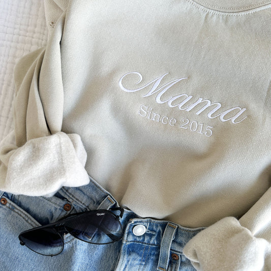 styled flat lay of a sand crewneck sweatshirt with embroidered Mama since customizable date in white thread across the chest with jeans and sunglasses