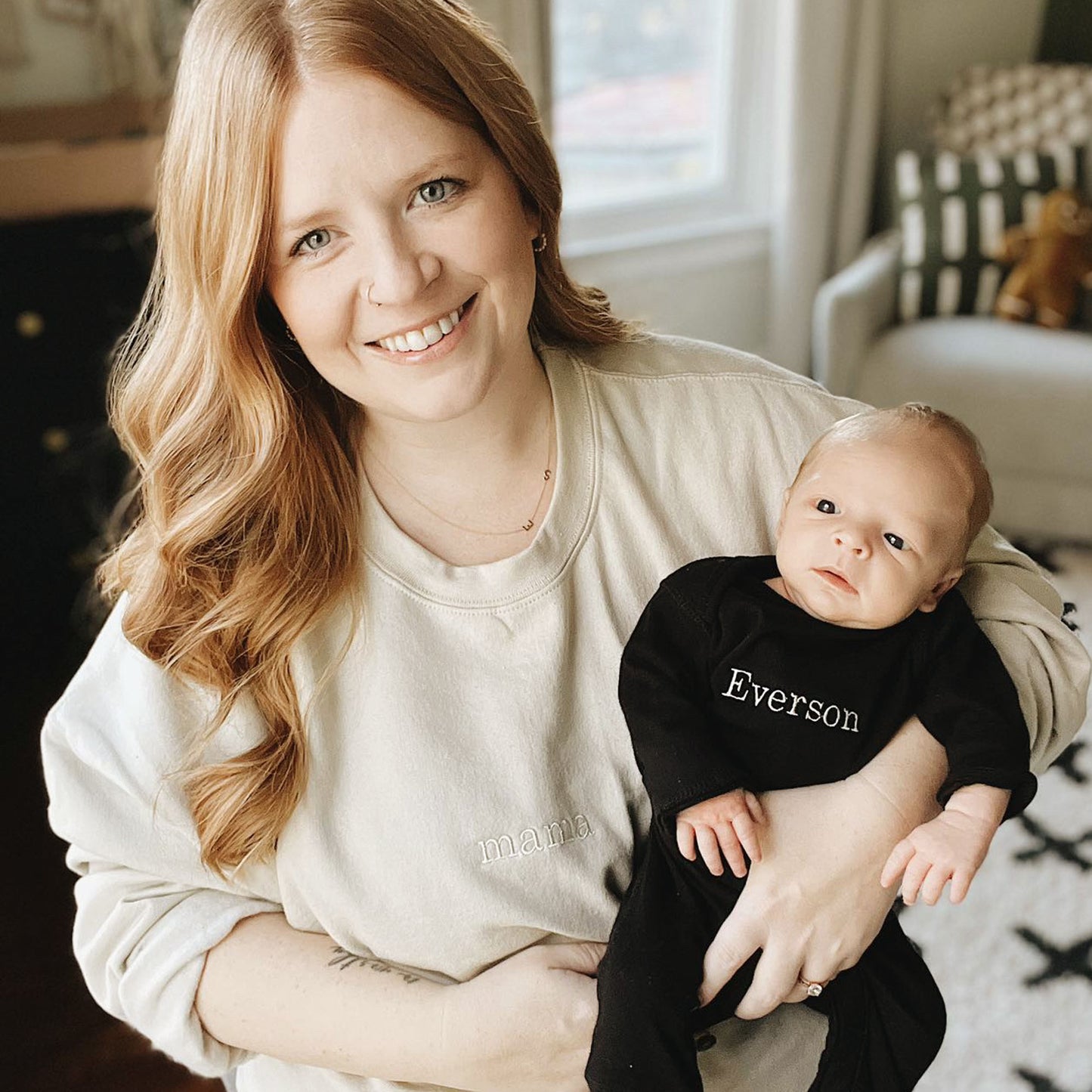 mom wearing a sand crewneck pullover with mama embroidered on the  center chest holding her baby who's wearing a long sleeve, long pant infant bodysuit with a custom name embroidered design