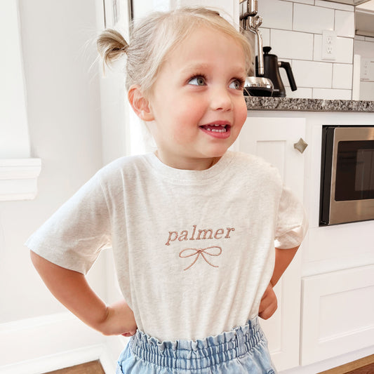 little girl wearing a heather oat t-shirt with custom name and dainty bow embroidered design on the center chest