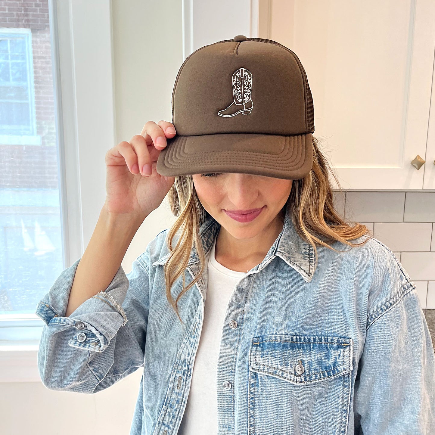 Woman wearing a brown trucker hat with embroidered cowgirl boot outline design in natural thread