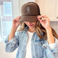 Woman wearing a brown trucker hat with embroidered cowgirl boot outline design in natural thread