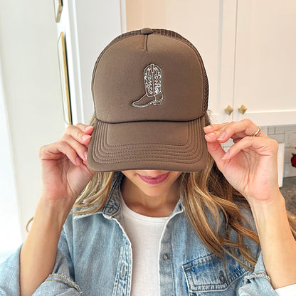 Woman wearing a brown trucker hat with embroidered cowgirl boot outline design in natural thread