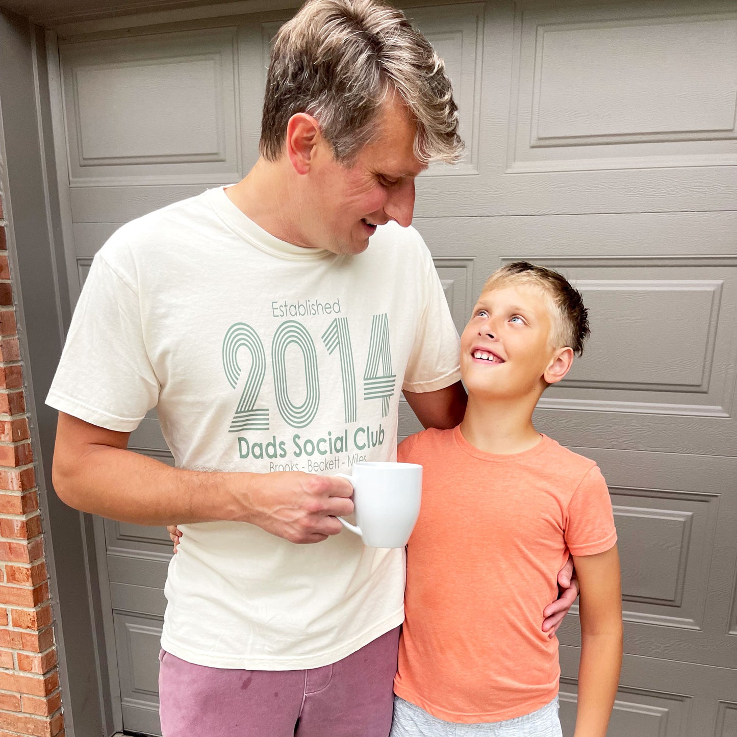 dad and son standing side-by-side. dad is wearing an ivory custom printed tee with his kids' names and date he became a dad