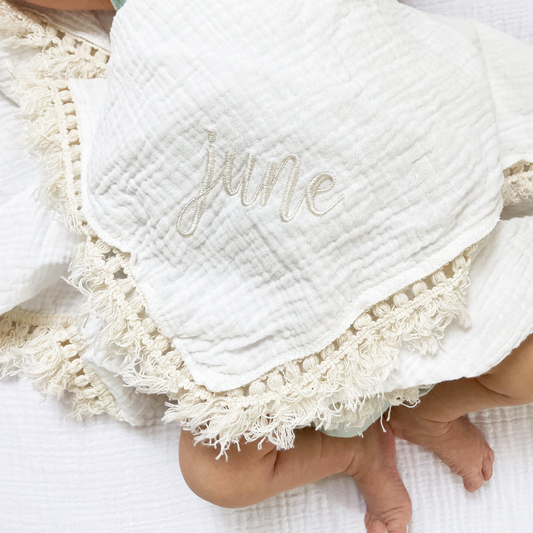 close up of a muslin baby blanket with decorative trim and custom embroidered name design on the corner 