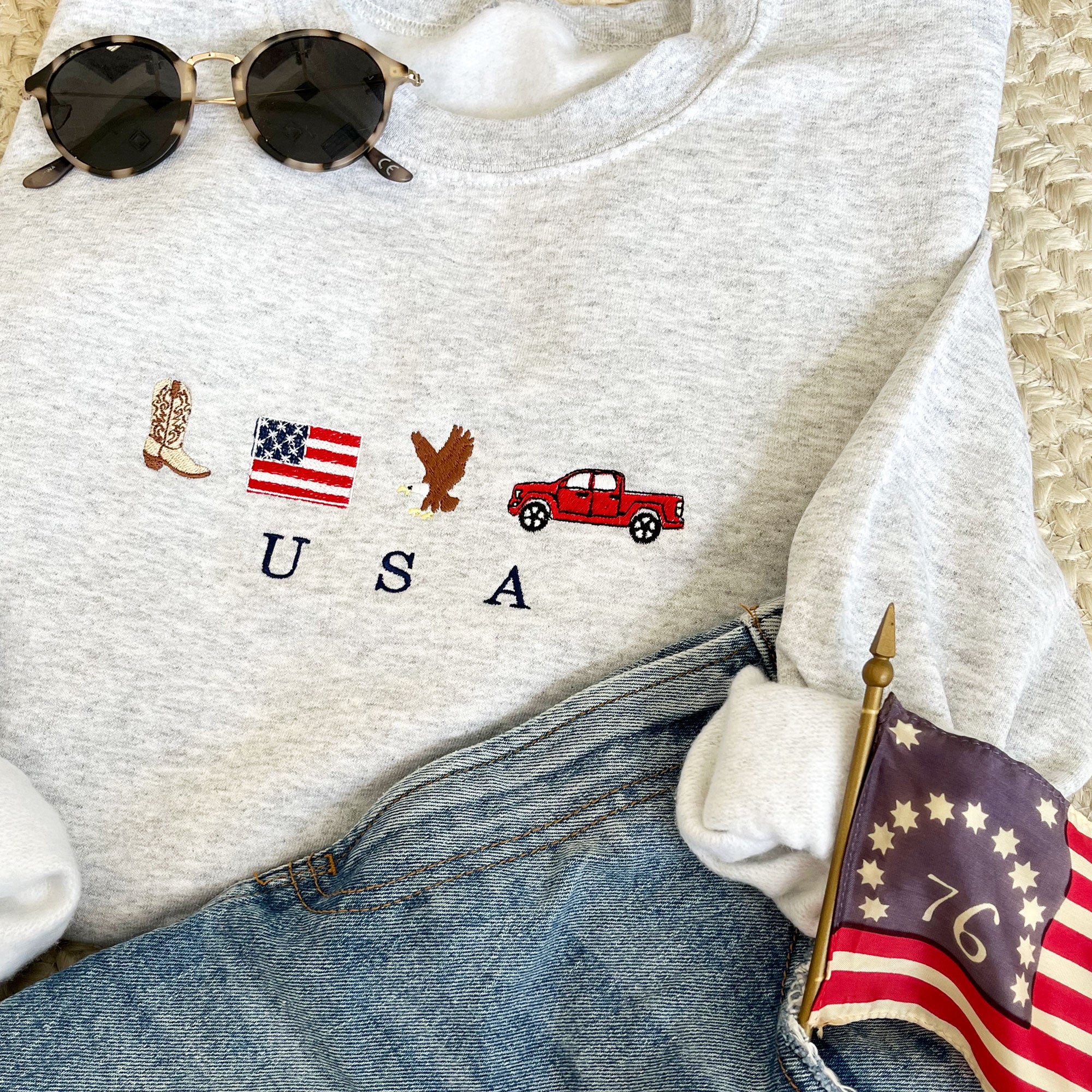 outfit layout featuring blue jean shorts, sunglasses, and an ash crewneck sweatshirt with cute USA icon embroidered design across the chest