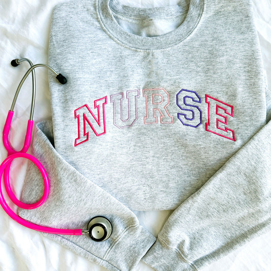 flat lay photo of an ash crewneck sweatshirt with embroidered athletic block nurse curved across the chest in alternating shades of pink and purple threads