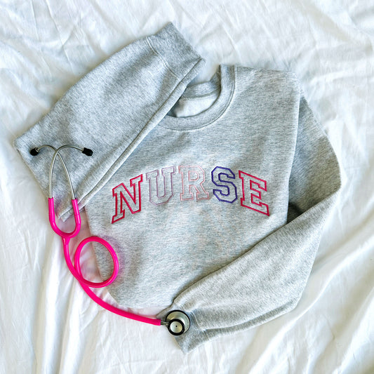 flat lay photo of an ash crewneck sweatshirt with embroidered athletic block nurse curved across the chest in alternating shades of pink and purple threads