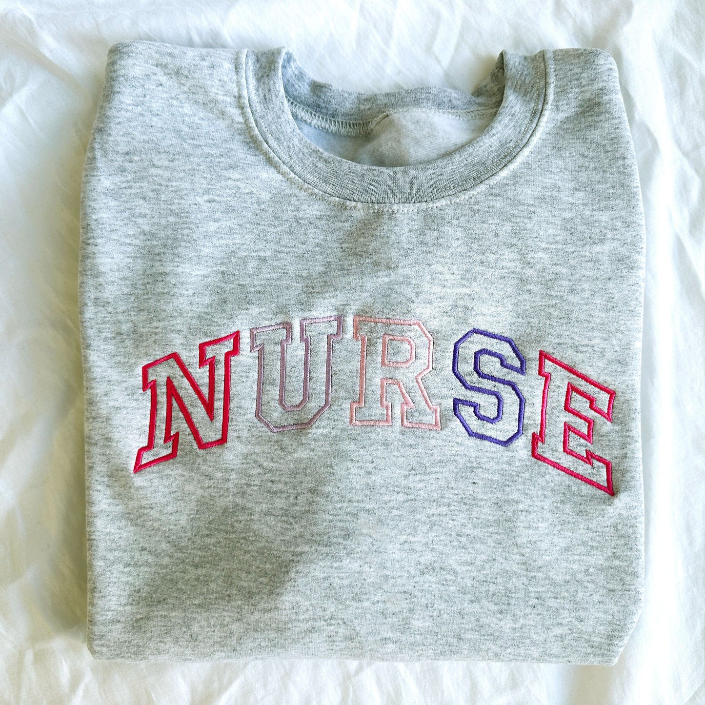 flat lay photo of an ash crewneck sweatshirt with embroidered athletic block nurse curved across the chest in alternating shades of pink and purple threads