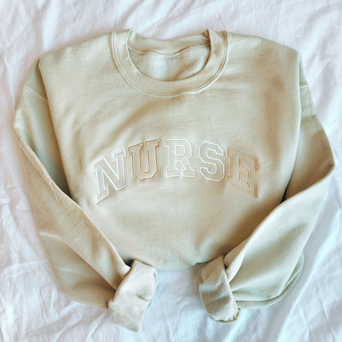 light sand crewneck pullover with NURSE embroidered across the chest in an athletic outline block font in neutral  thread colors