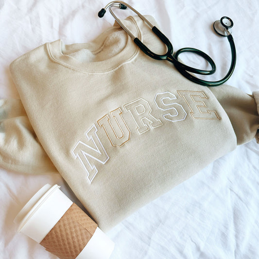 coffee cup, stethoscope, and sand crewneck sweatshirt with NURSE embroidered across the chest in neutral thread colors