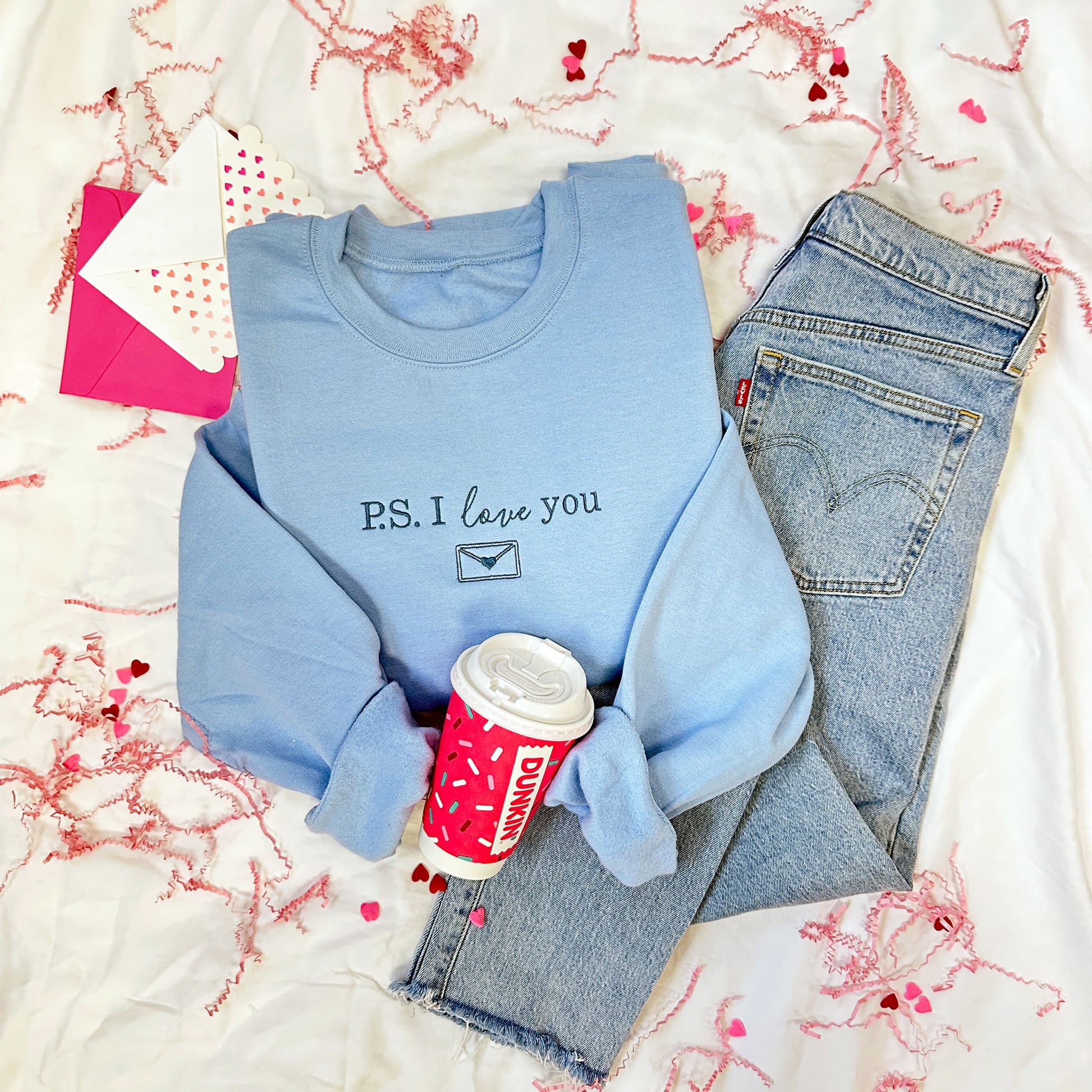 flat lay image of jeans, dunkin' coffee cup, and a blue crewneck sweatshirt with a P.S. I love you design embroidered across the chest