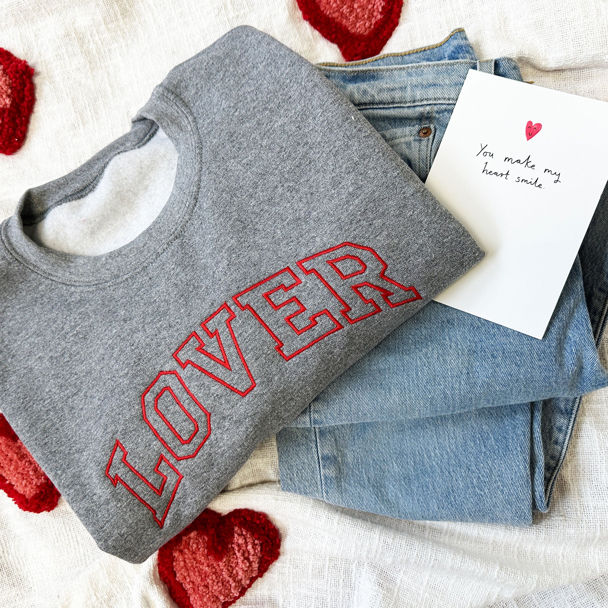heart blanket, cute valentine's gift card, jeans, and a gray sweatshirt with an open block athletic block font reading LOVER embroidered across the chest in red