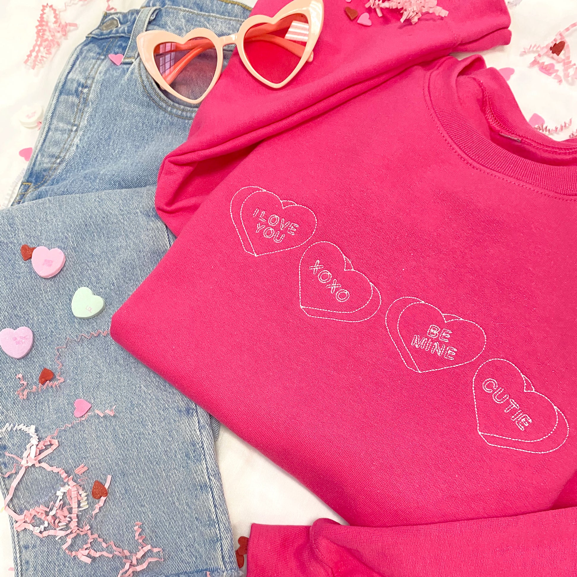 flat lay image of jeans, heart shaped glasses, a bright pink crewneck sweatshirt with stitched candy hearts across the chest  with "I love you," "xoxo," "be mine," and "cutie" designs
