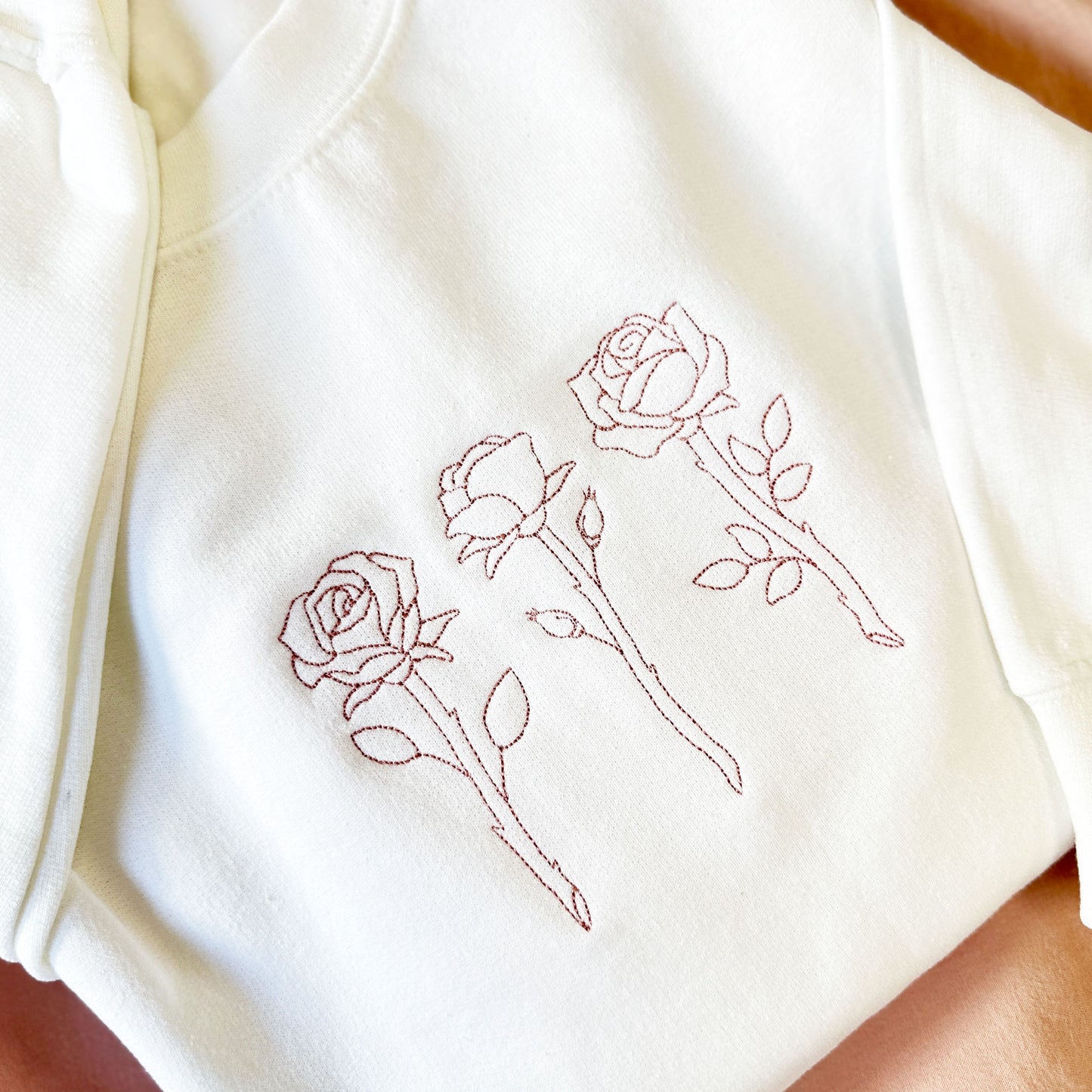 close up of a white crewneck sweatshirt with  stitched roses embroidered across the chest in a mauve thread.