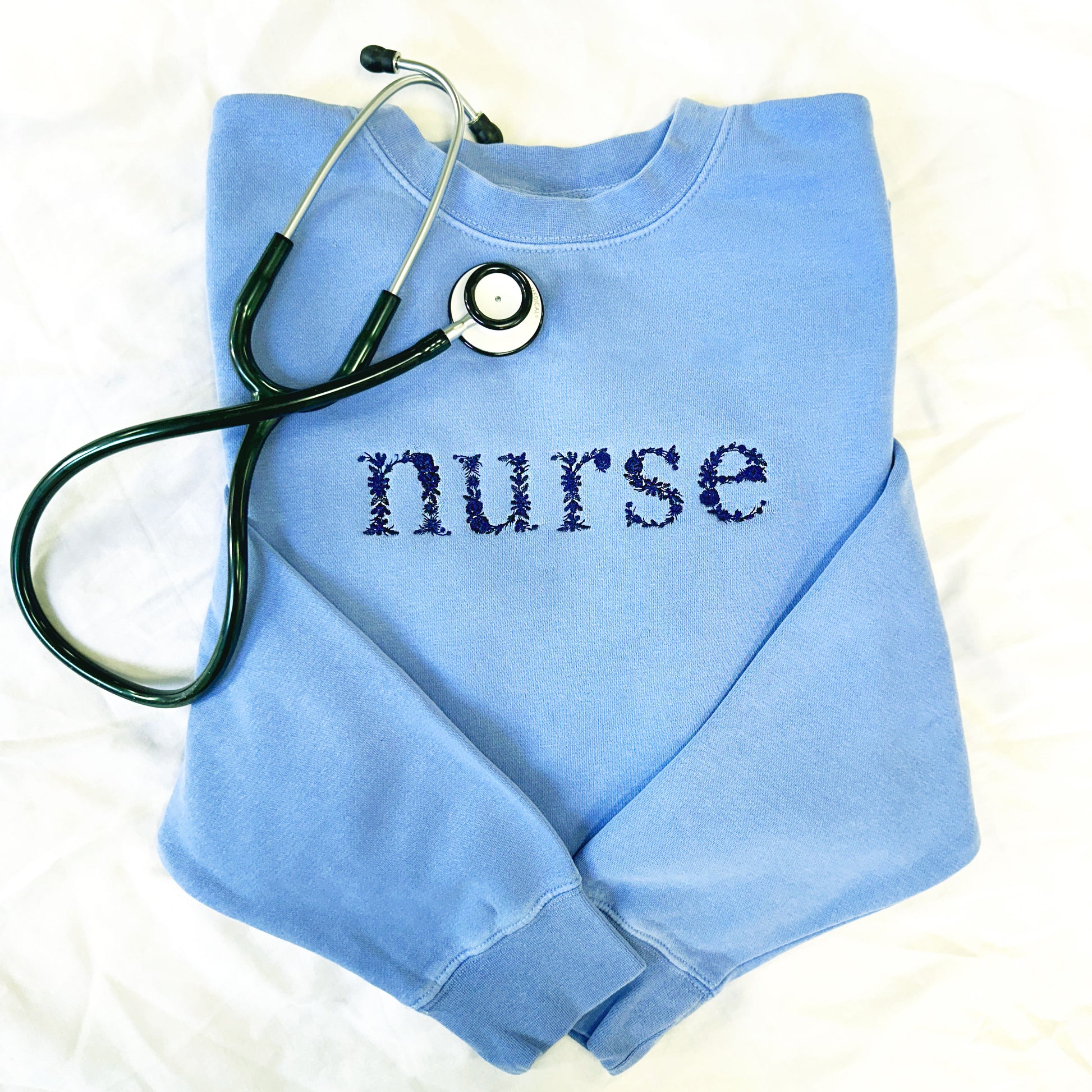 Flat lay of an embroidered nurse light blue crewneck sweatshirt with floral letters in navy