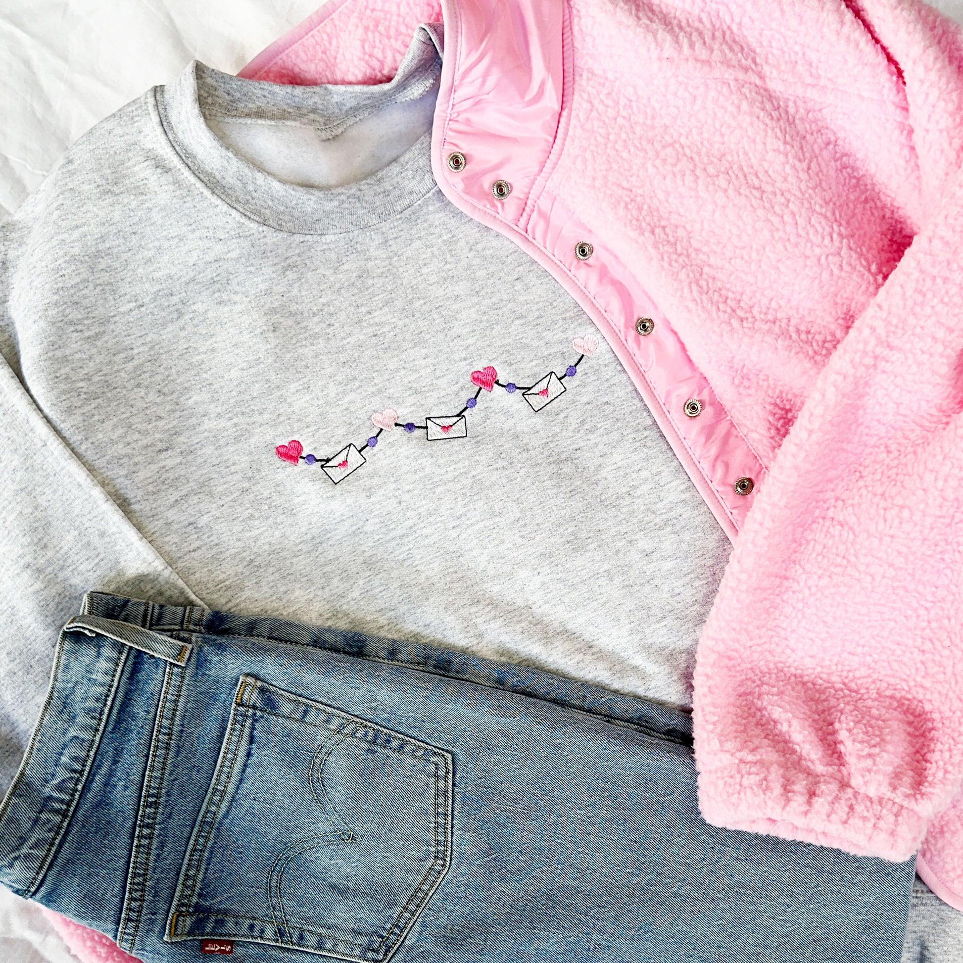 jeans and a bright pink fleece jacket paired with a light gray sweatshirt with a valentine's themed garland design embroidered on the center chest featuring mini hearts and mini envelopes
