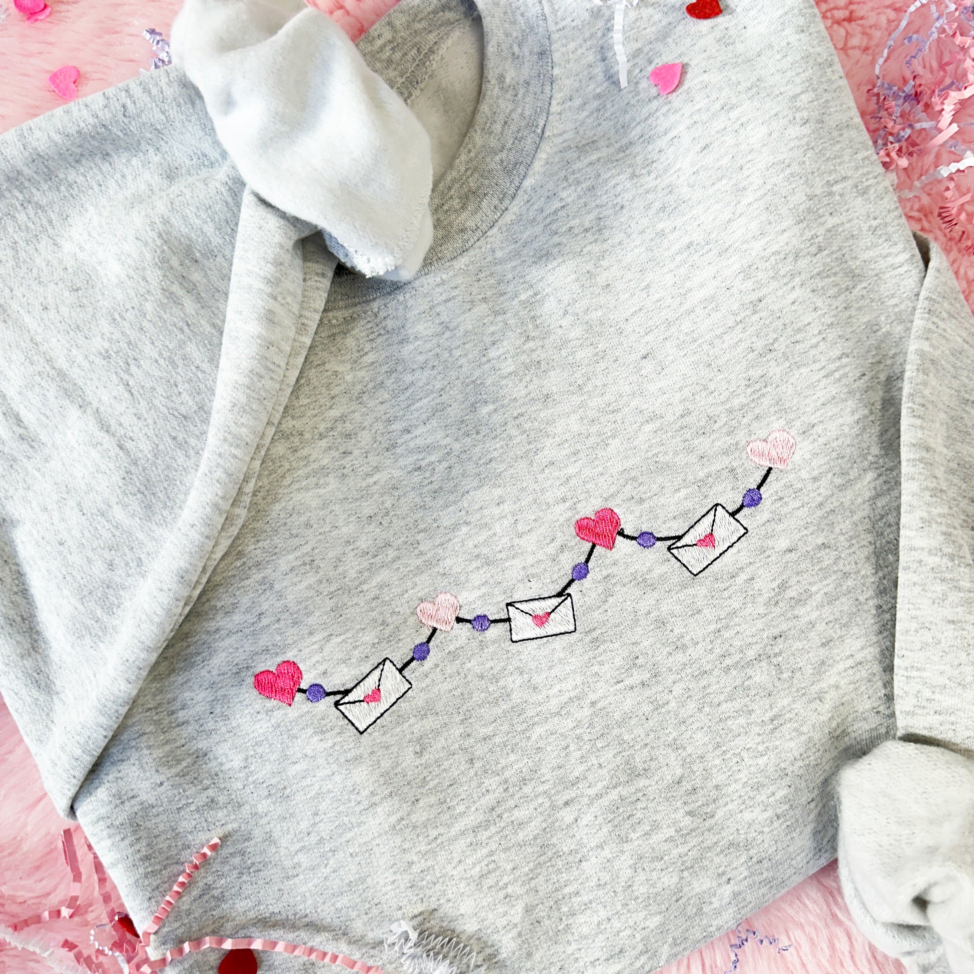 close up image of an ash gray crewneck sweatshirt with a valentine's pink and purple garland design across the center chest