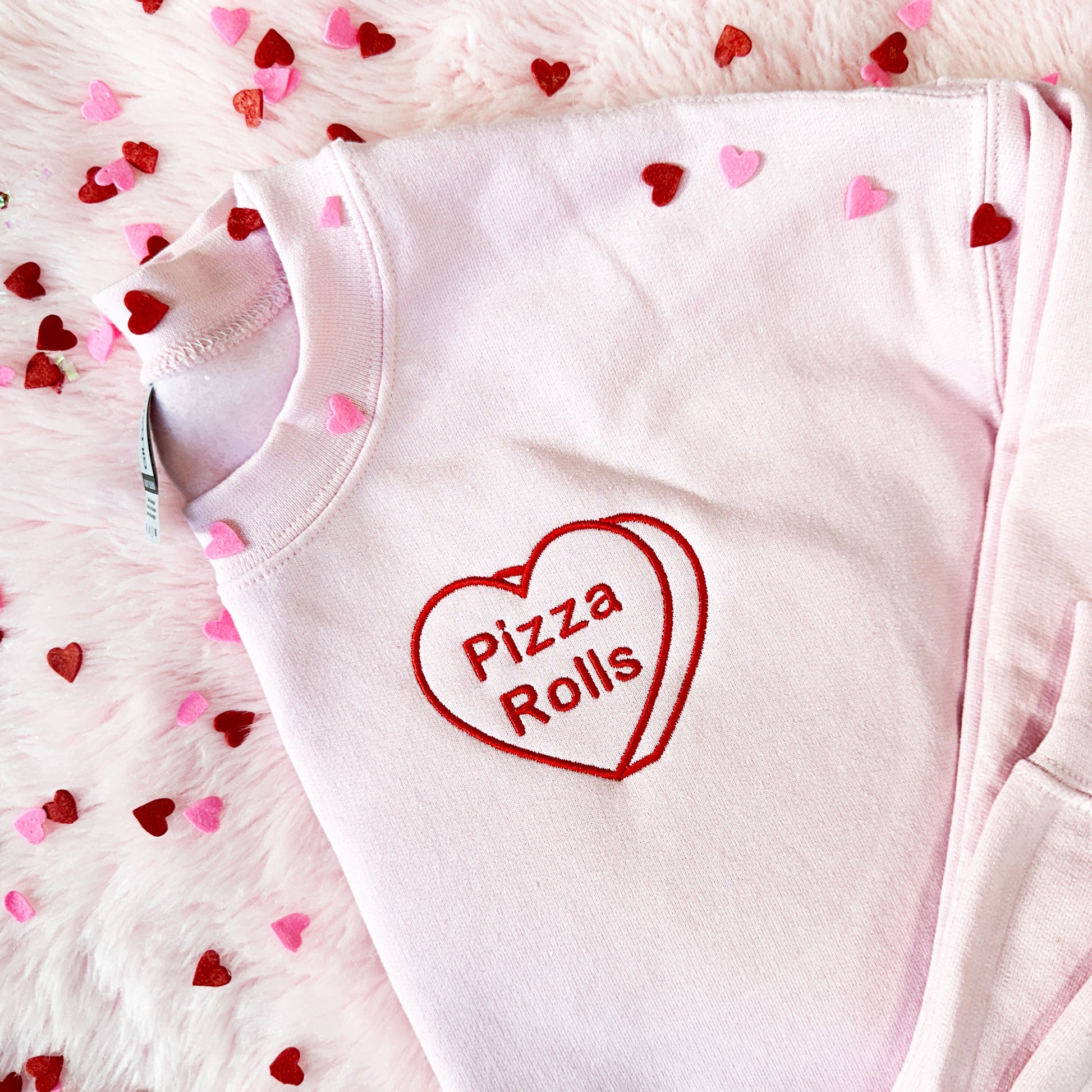 Flat lay image of a pink crewneck sweatshirt with a candy heart embroidered design that says pizza rolls in red thread 