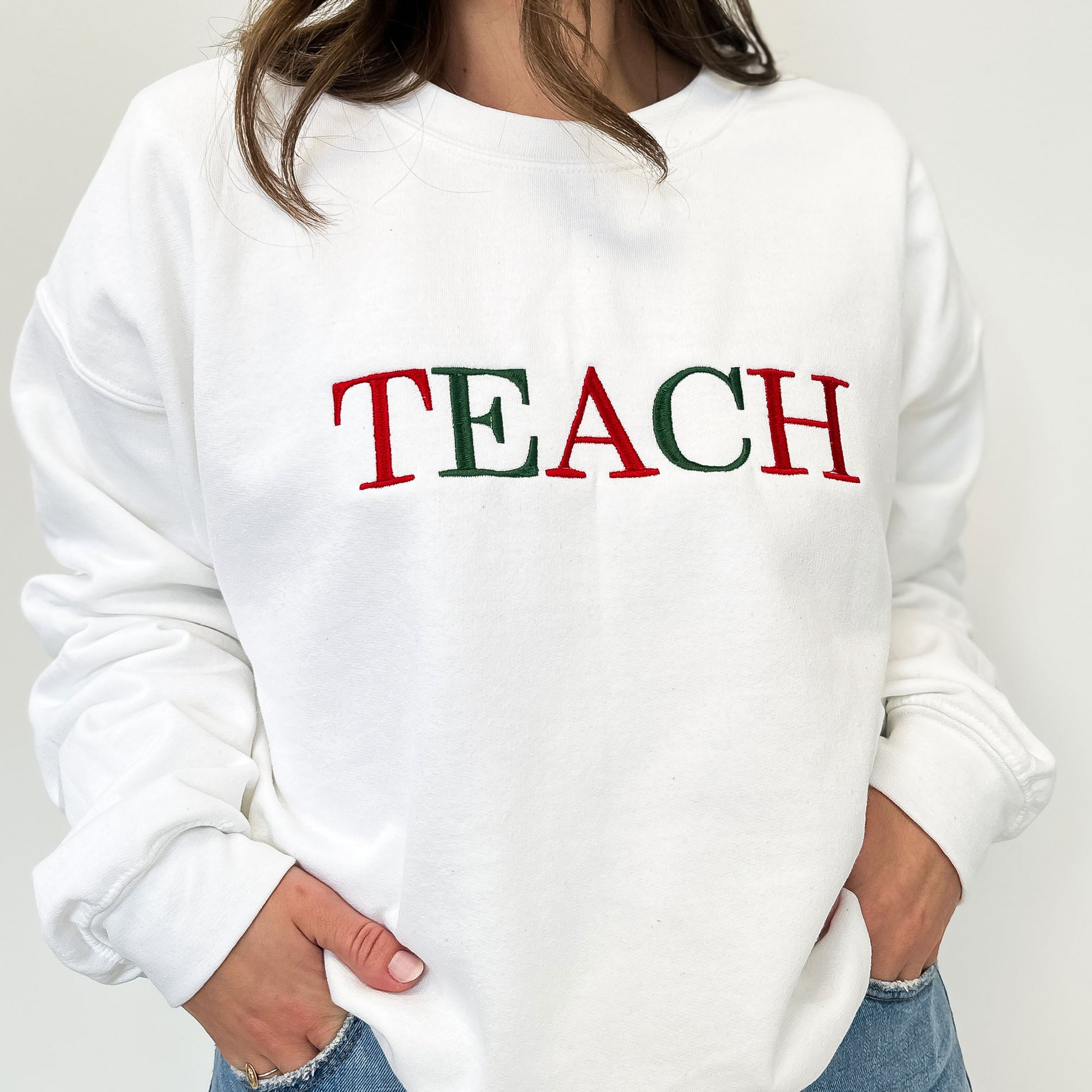 Close up of a woman wearing a white crewneck sweatshirt that has Teach embroidered in all uppercase letter in alternating red and green threads.