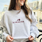 Close up of woman wearing an ash crewneck sweatshirt with rose design in la vie en rose text embroidered in burgundy across the center chest