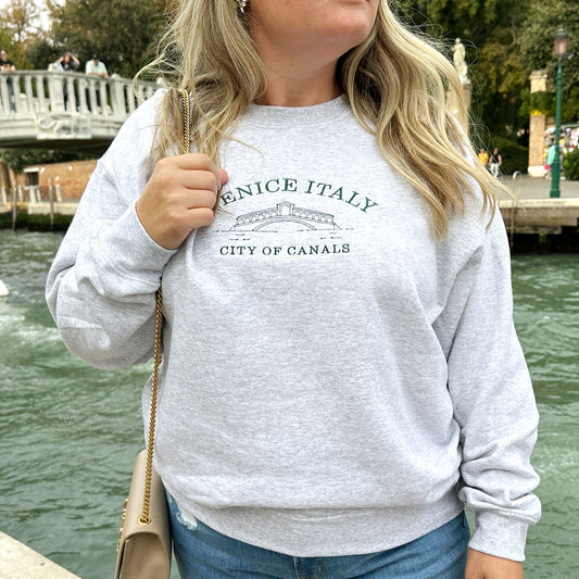Woman wearing an ash crewneck sweatshirt with Venice Italy city of canals and sketch of bridge embroidered in ivy across the chest