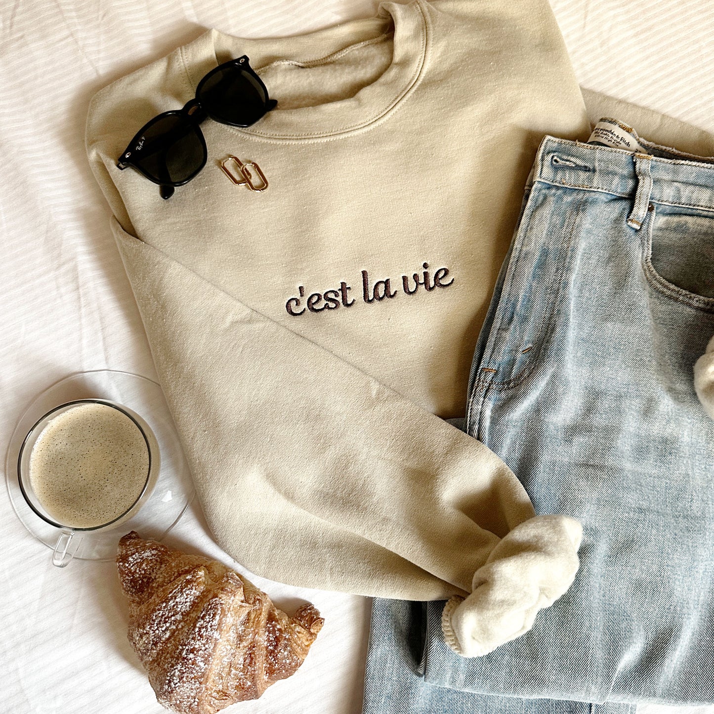 styled flat lay of c'est la vie sand crewneck embroidered sweatshirt with jeans, sunglasses, coffee and croissant