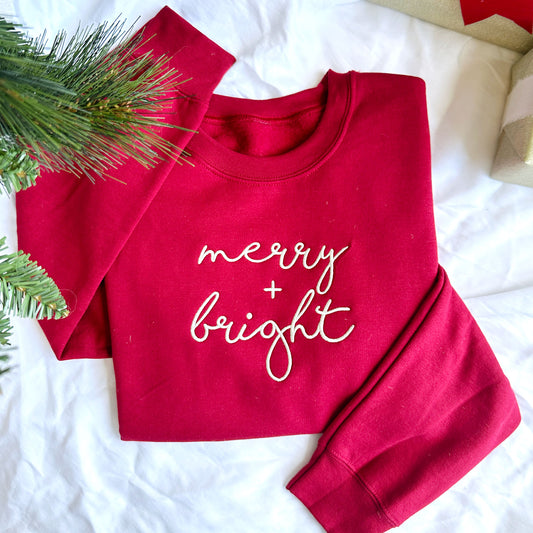 red crewneck sweatshirt with a merry and bright embroidered design on the chest laying under a christmas tree