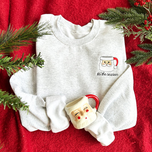 Flat lay image of an ash crewneck sweatshirt laying on a red blanket. the sweatshirt has embroidered on the left chest a santa mug design and the words tis the season below in cursive.