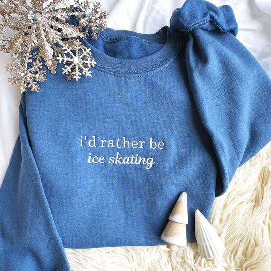 festive flat lay image of an indigo crewneck sweatshirt with i'd rather be ice skating embroidered across the chest