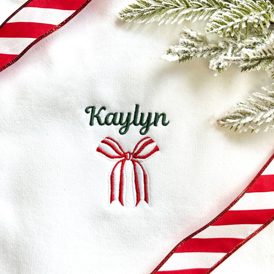 close up image of an embroidered name and festive holiday ribbon bow on a white sweatshirt