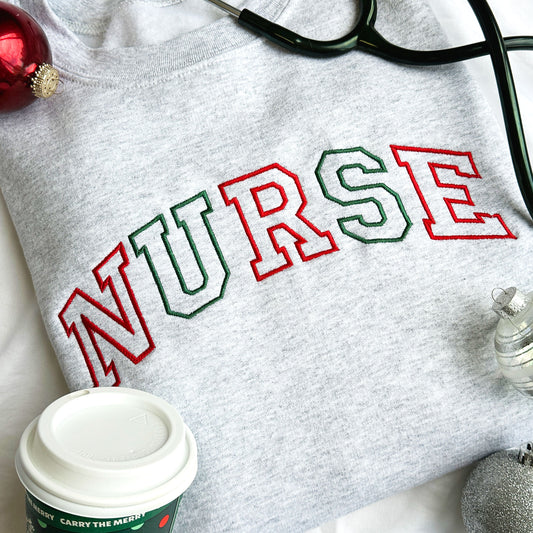 Close up flat lay image of an ash crewneck sweatshirt featuring the word Nurse embroidered in red and green across the chest.