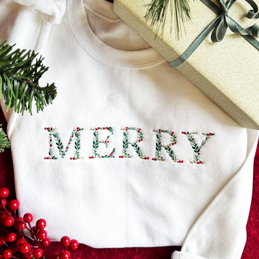 Flat lay image of a white crewneck sweatshirt with the word MERRY embroidered in floral text with red, gold and green colors.