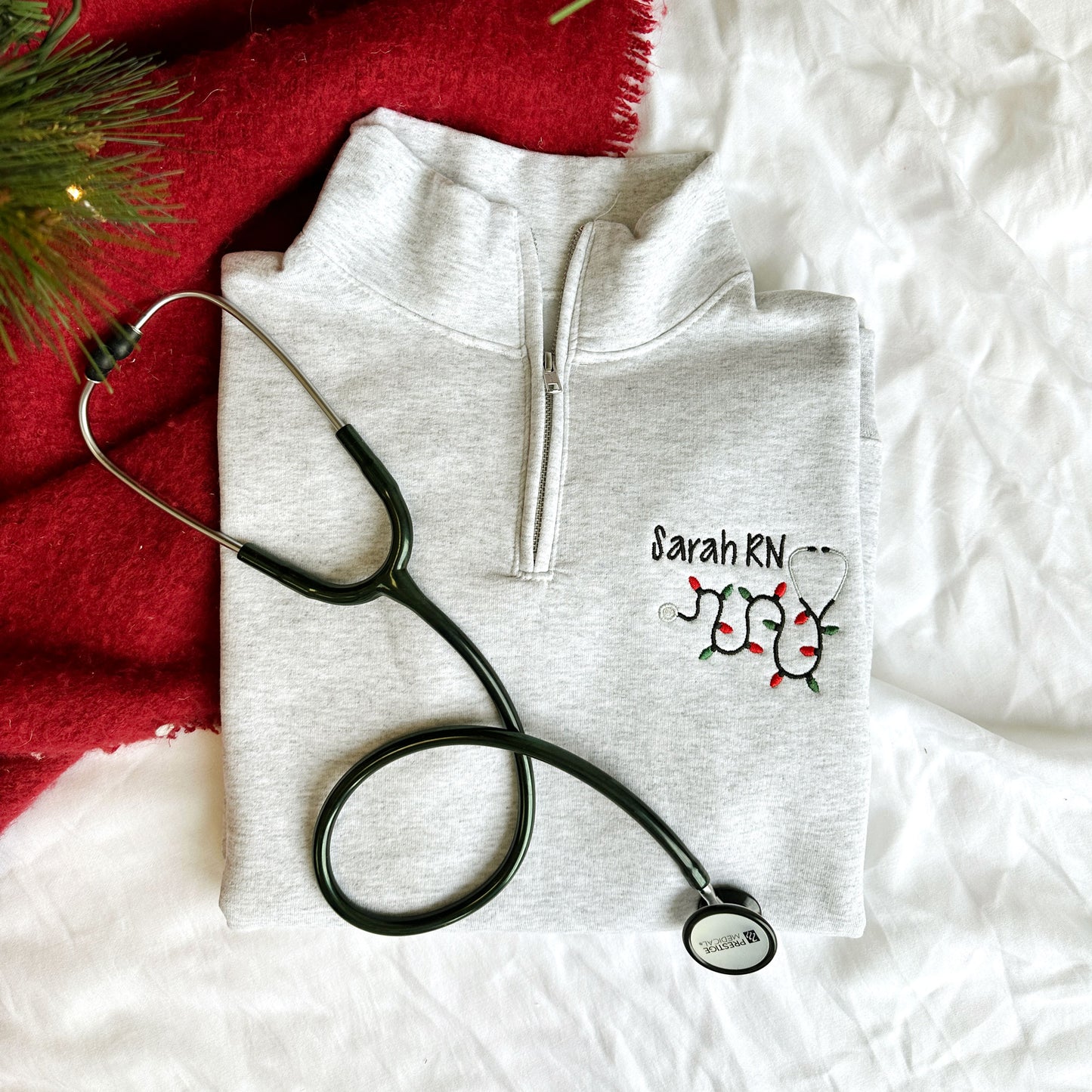 flat lay image of a stethoscope and a light gray quarter zip sweatshirt with a custom name and christmas lights stethoscope embroidery on the left chest