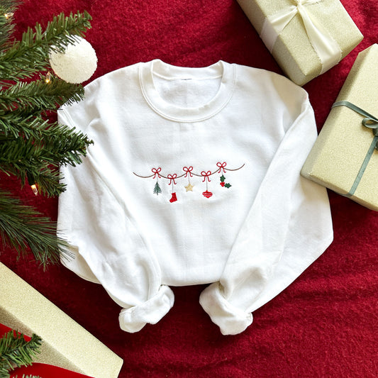 Flat lay image of a white crewneck sweatshirt with embroidered traditional Christmas garland. The garland features a christmas tree, stocking, star, ornament, and holly leaf.