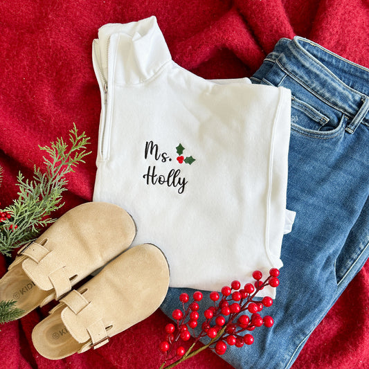 flat lay of a teacher outfit featuring birkinstock clogs, blue jeans, and a white quarter zip with a custom teacher name and holly embroidery design