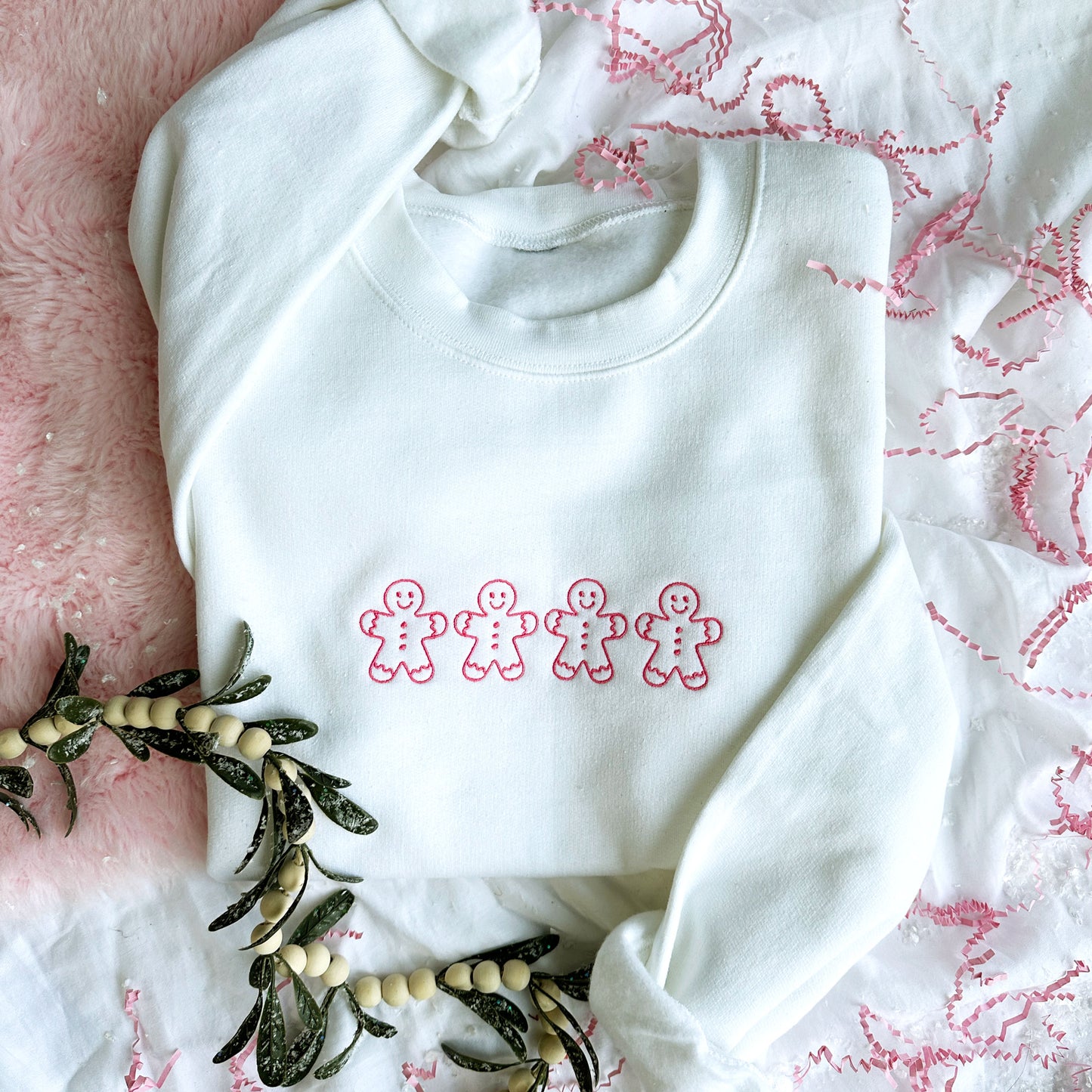 Cute styled flat lay of a white crewneck sweatshirt featuring 4 stitched outline gingerbread men embroidered in pink. 