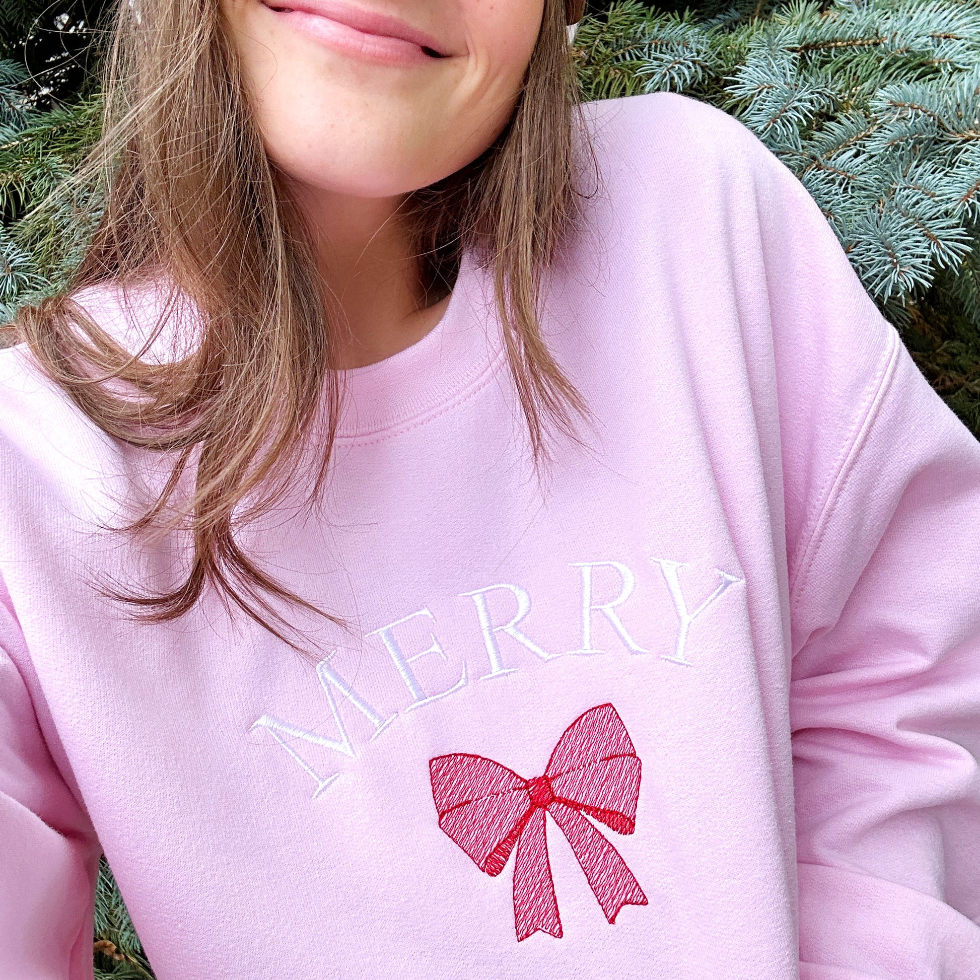 Close up image of a woman wearing a light pink crewneck sweatshirt showcasing the embroidered the design. The embroidery features the word Merry in all capital letters arched above a stitched red bow.