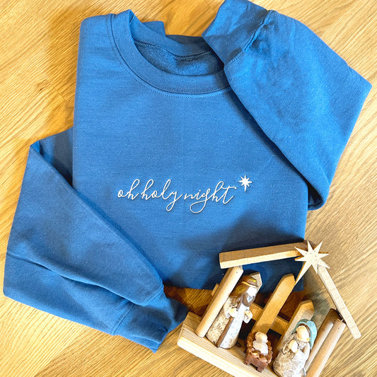 flat lay image of a blue sweatshirt with oh holy night embroidered in a script font across the chest with a small wooden nativity laying near it