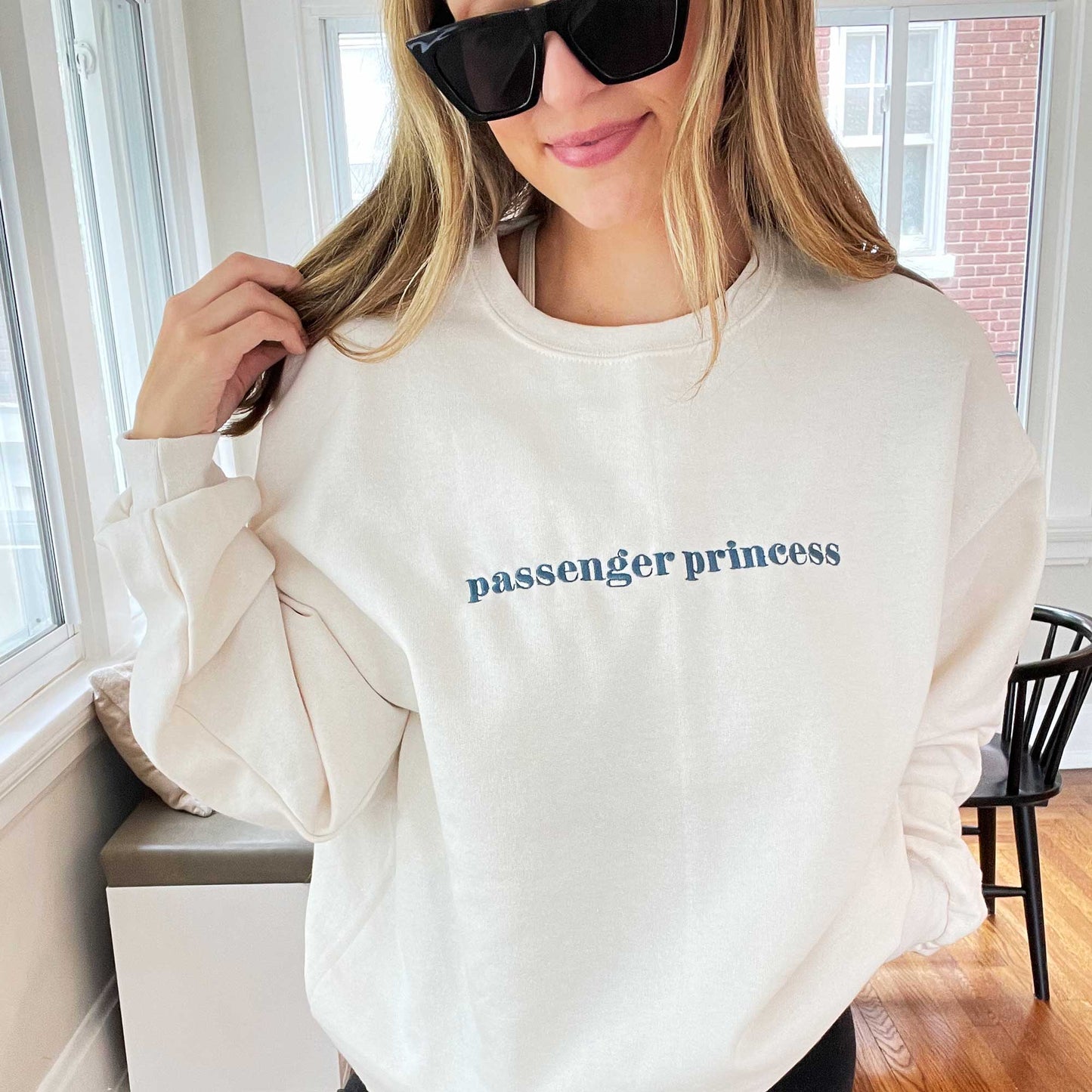 young woman wearing sunglasses and a sweet cream crewneck sweatshirt with passenger princess embroidered in french blue thread