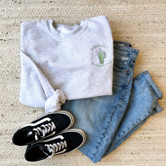 flat lay layout of an outfit featuring vans, jeans, and a crewneck sweatshirt with a stanley cup embroidered design on the left chest.