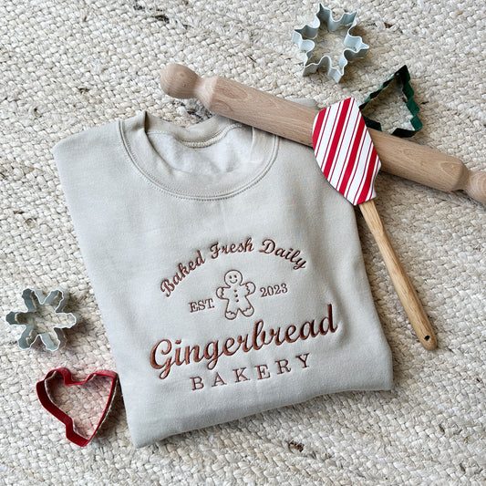 flat lay image of cookie cutters and a rolling pin surrounding a light brown sweatshirt with a festive "baked fresh daily Gingerbread Bakery" design embroidered across the chest