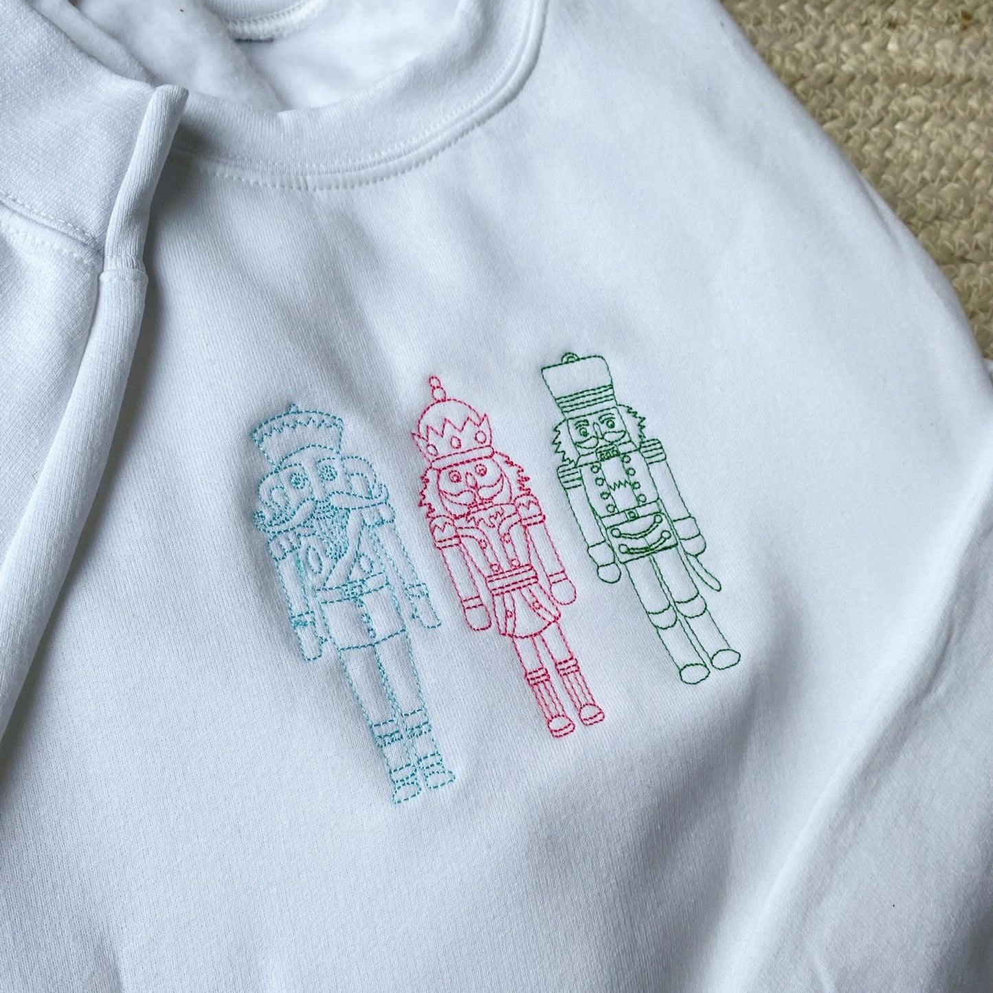 Up close image of a white crewneck sweatshirt featuring stitched embroidered nutcrackers across the chest in blue, pink, and green threads.