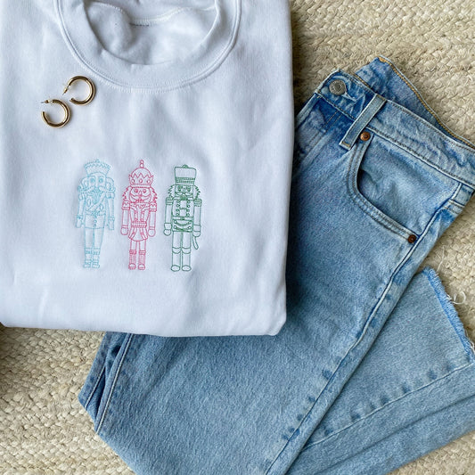 Styled flat lay image of a white crewneck sweatshirt and blue jeans. the white crewneck features 3 stitched nutcrackers in blue, pink, and green threads.