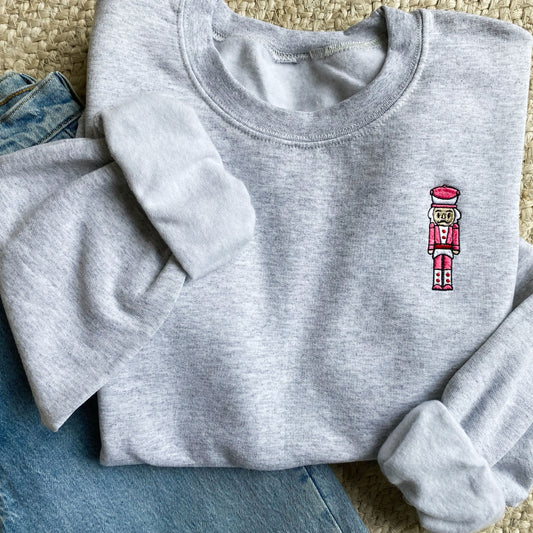 Flat lay of an ash crewneck sweatshirt and a pair of blue jeans. The sweatshirt features a small embroidered pink and red nutcracker on the left chest.