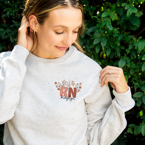 woman standing outside wearing an ash gray sweatshirt featuring a custom RN and name fall floral print on the left upper chest