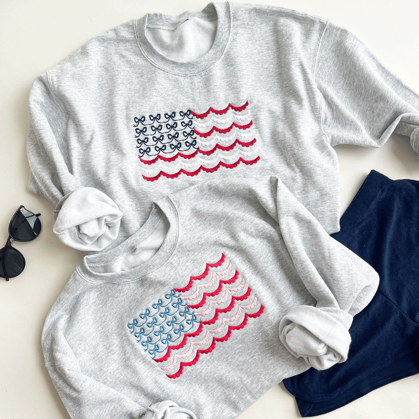 May include: Two gray sweatshirts with embroidered American flags. The flags are made of blue bows and red, white, and pink scalloped edges.