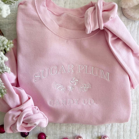 Flat lay image of a light pink crewneck sweatshirt featuring Sugar Plum Candy Co embroidered in white thread across the chest.