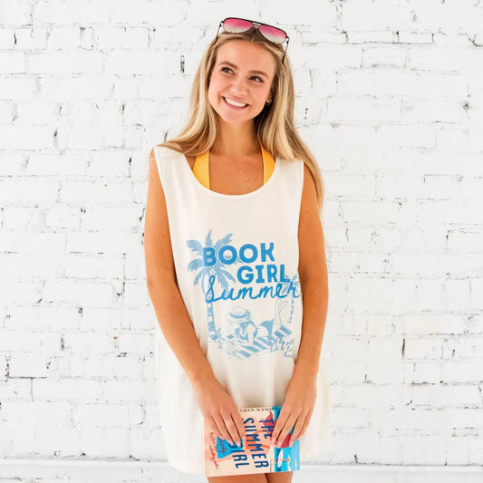 girl standing in front of a white brick wall holding a book and wearing an oversized comfort colors tank top over a bathing suit like a  cover up. The tank top has a book girl summer print in blue ink featuring a palm tree, and a girl reading and laying out on a beach towel