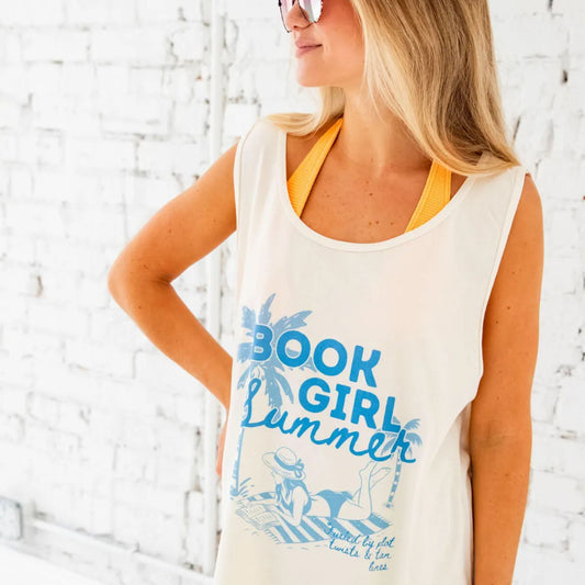 close up of a girl standing in front of a white brick wall holding a book and wearing an oversized comfort colors tank top over a bathing suit like a  cover up. The tank top has a book girl summer print in blue ink featuring a palm tree, and a girl reading and laying out on a beach towel