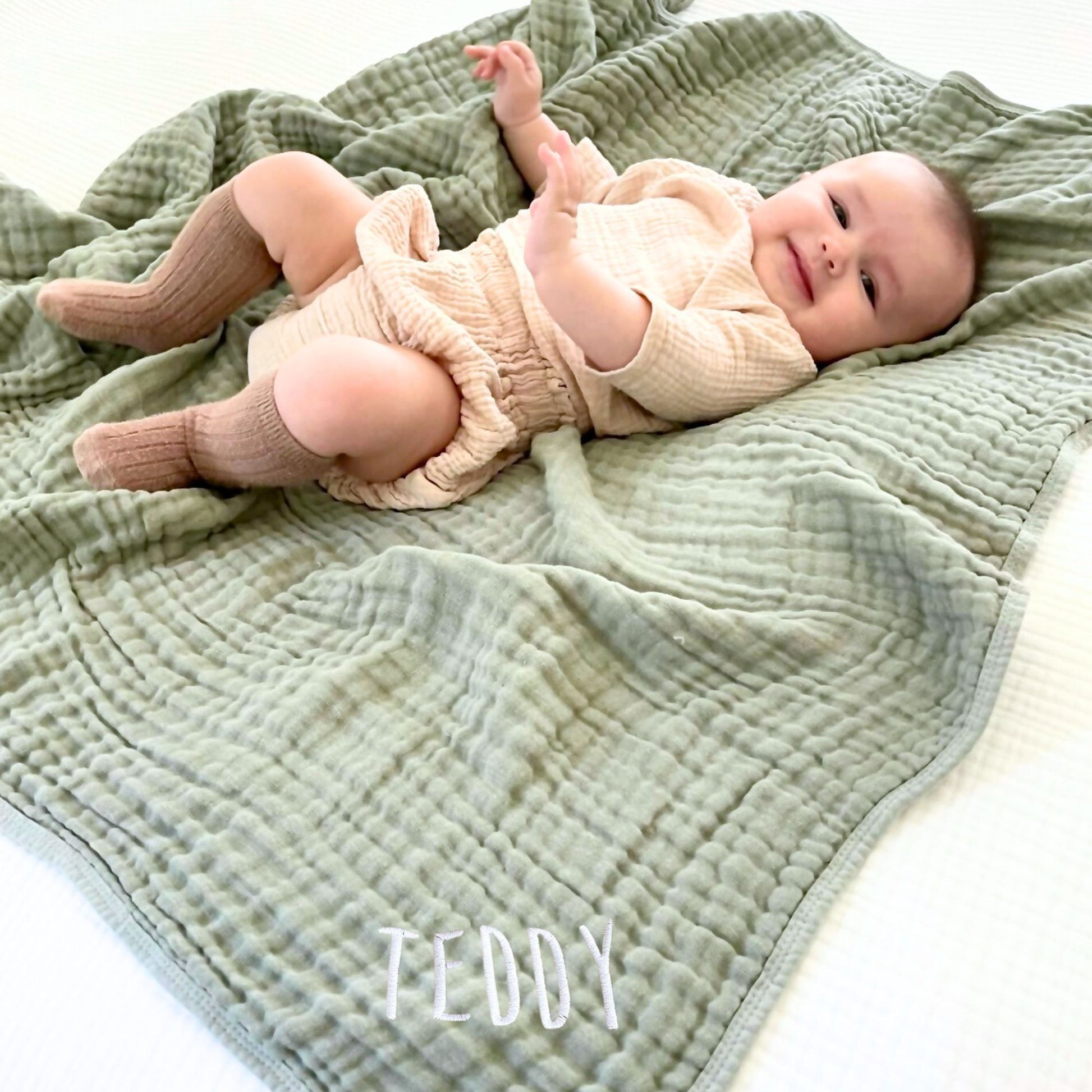 baby laying on a fern green muslin blanket with custom name embroidery in the corner